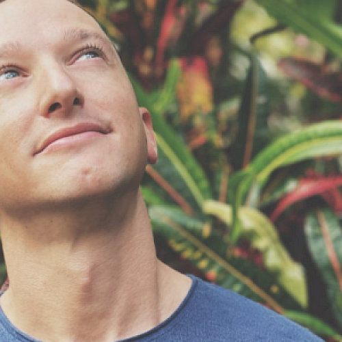 A person in a blue shirt looks upwards while standing in front of colorful foliage in a natural setting.