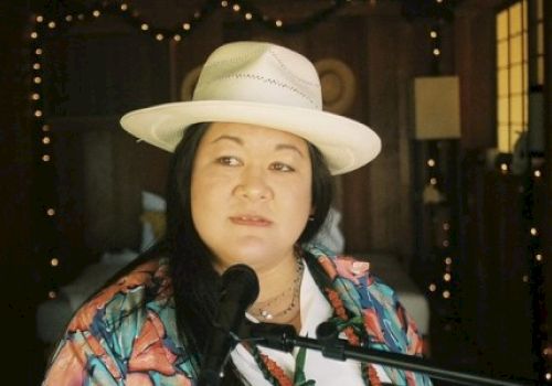 A person is wearing a hat and colorful shirt while speaking or singing into a microphone. The background features string lights and a warmly lit room.