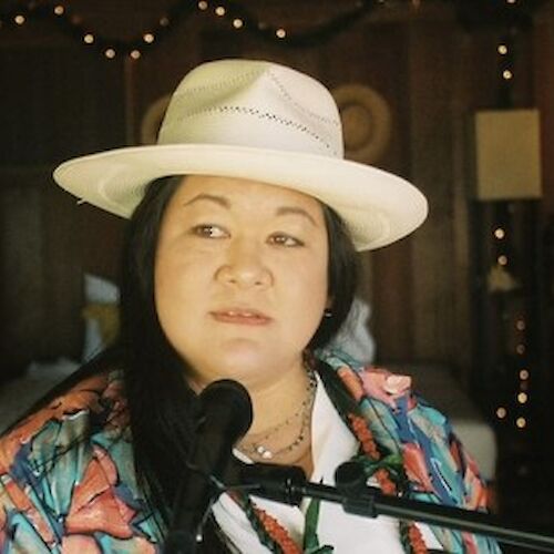 A person is wearing a hat and colorful shirt while speaking or singing into a microphone. The background features string lights and a warmly lit room.