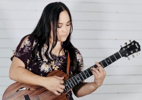 A person is playing an acoustic guitar against a white, horizontally slatted background.