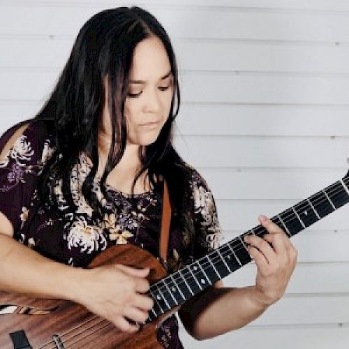 A person is playing an acoustic guitar against a white, horizontally slatted background.