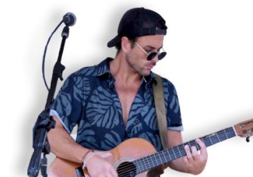 A person in sunglasses and a hat is playing an acoustic guitar next to a microphone stand, wearing a patterned shirt, with a white background.