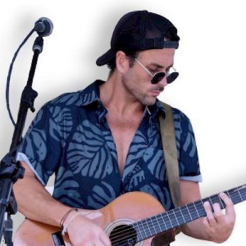 A person in sunglasses and a hat is playing an acoustic guitar next to a microphone stand, wearing a patterned shirt, with a white background.