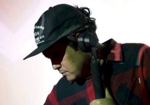 A person wearing a black cap and a red and black checkered shirt is DJing with headphones on against a light-colored background.