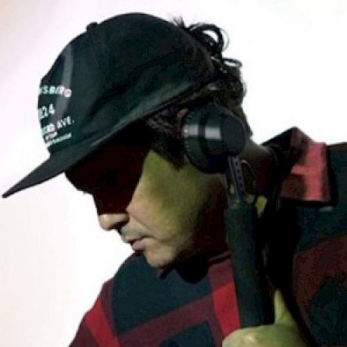 A person wearing a black cap and a red and black checkered shirt is DJing with headphones on against a light-colored background.