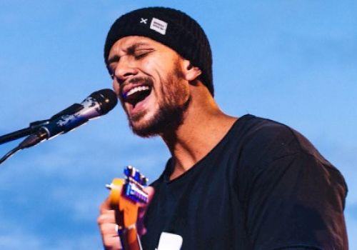 A bearded man wearing a beanie and black shirt passionately sings into a microphone while playing a ukulele, set against a clear blue sky.