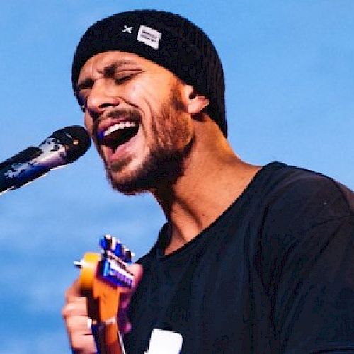 A bearded man wearing a beanie and black shirt passionately sings into a microphone while playing a ukulele, set against a clear blue sky.