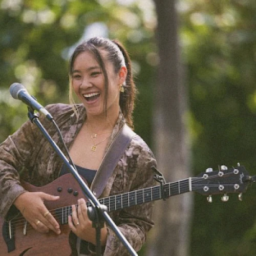 Una mujer sonriente con una guitarra est&aacute; de pie junto a un micr&oacute;fono, actuando al aire libre con vegetaci&oacute;n de fondo.