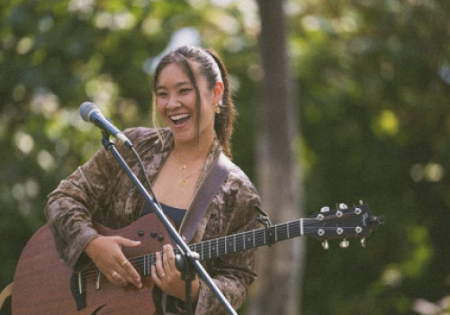 A smiling woman with a guitar stands next to a microphone, performing outdoors with greenery in the background.