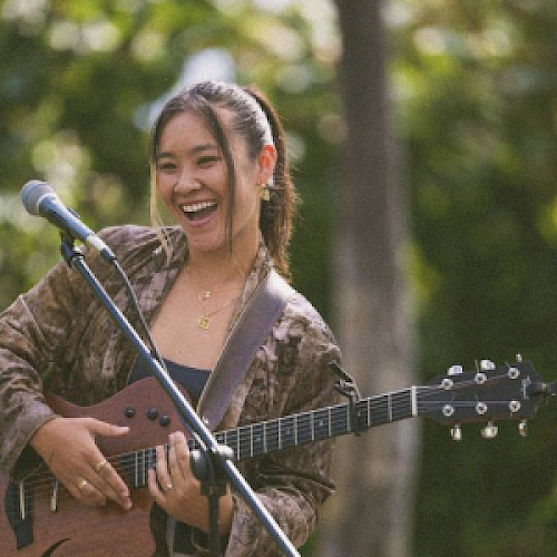 A smiling woman with a guitar stands next to a microphone, performing outdoors with greenery in the background.