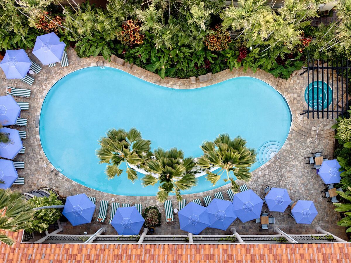 Aerial view of a kidney-shaped pool surrounded by palm trees, blue umbrellas, and lounge chairs; a small hot tub is also visible.