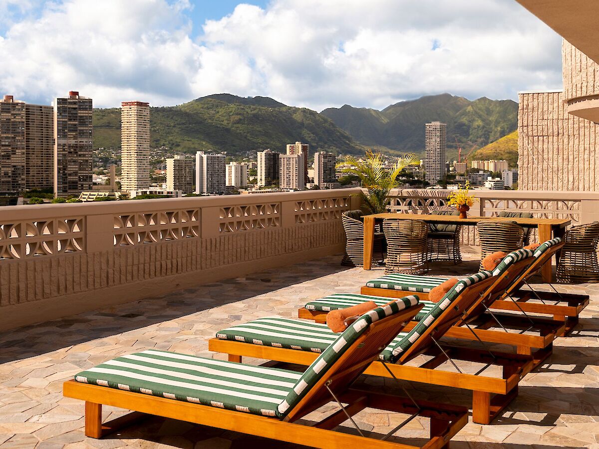 Outdoor terrace with reclining chairs, cityscape view, tall buildings, greenery, and mountains in the background under a partly cloudy sky.