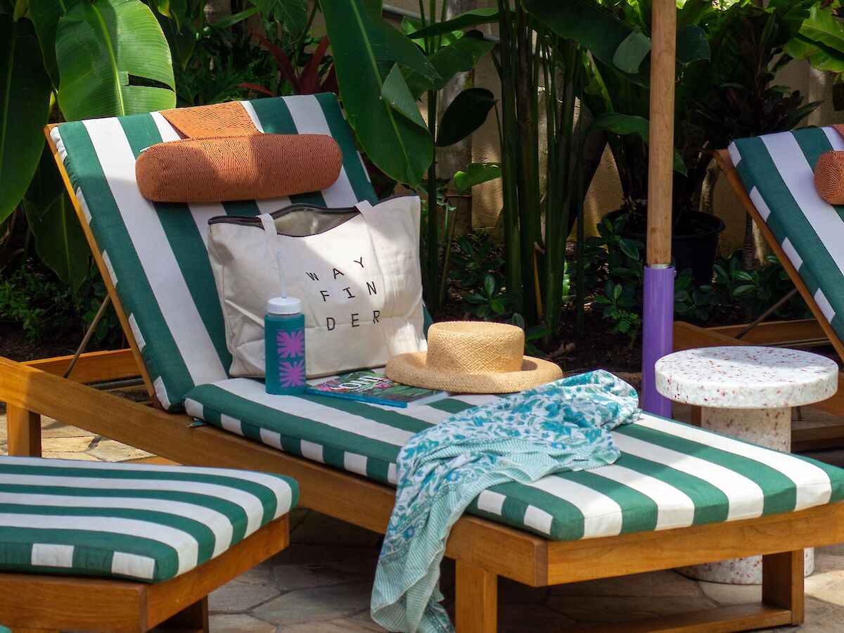 The image shows a cozy outdoor setup with striped lounge chairs, a hat, a water bottle, and a blue umbrella, surrounded by lush greenery.