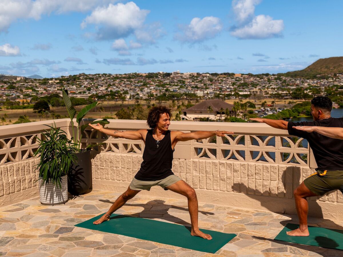 Two people practice yoga on a terrace overlooking a scenic view, performing a Warrior II pose on yoga mats, with plants and a city landscape in the background.