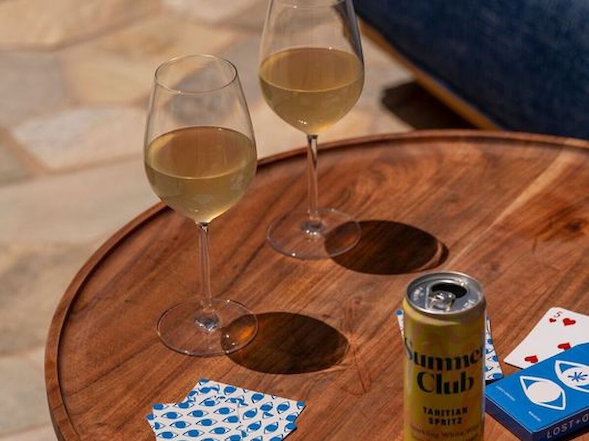 A wooden table with two glasses of white wine, a beer can, a paper napkin with blue dots, and a deck of cards.