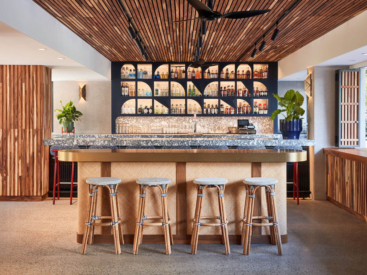 A modern bar with wooden stools, a granite countertop, and a backlit shelf featuring various bottles. The design includes plants and earthy tones.