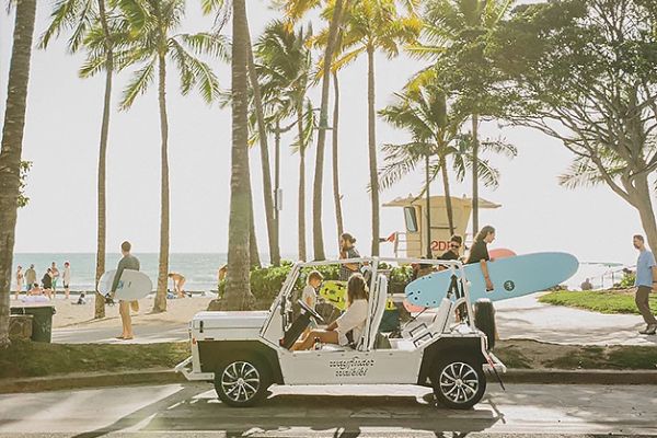 A beach scene with tall palm trees, people carrying surfboards, and a group of friends in a cart carrying a surfboard, enjoying the sunny day.