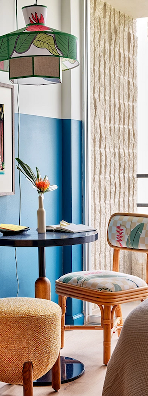 A small table with a notebook, pencil, vase with flower, mug, chair with cushions, and framed art on a blue wall.