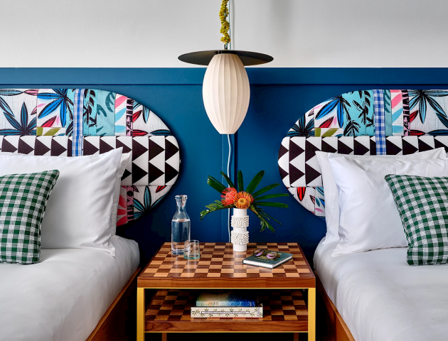 A hotel room features two double beds with colorful headboards, green checkered pillows, a side table with a lamp, flowers, and a hat.