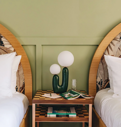 The image shows a cozy bedroom with two single beds, a green cactus-shaped lamp on a nightstand between them, and patterned headboards.