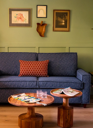 The image shows a cozy living space with a blue sofa, a small coffee table, framed pictures on the wall, a floor lamp, and a bed in the background.