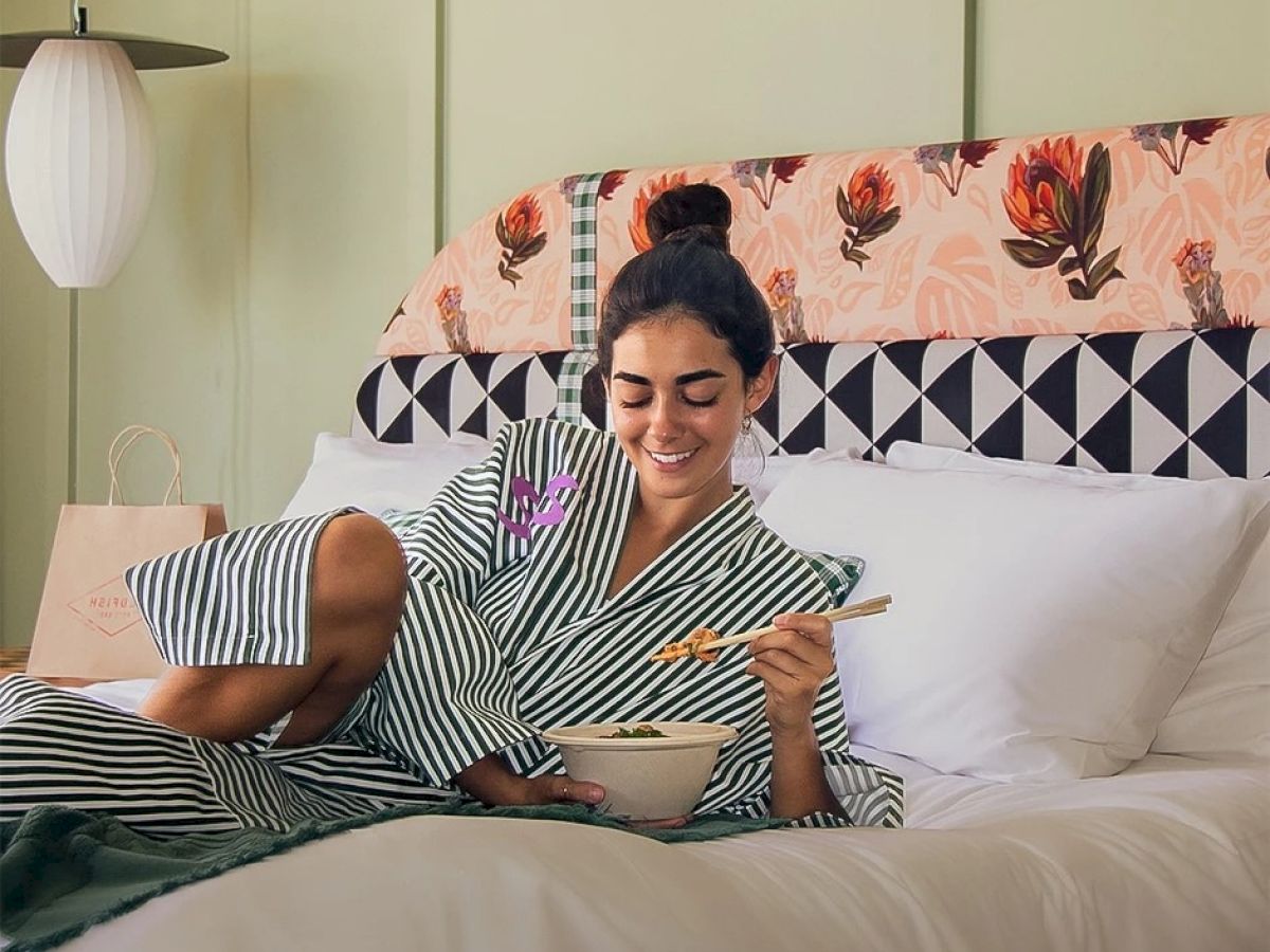 A person in a striped robe is sitting on a bed, eating with chopsticks from a bowl, and smiling. The bed has a colorful headboard.