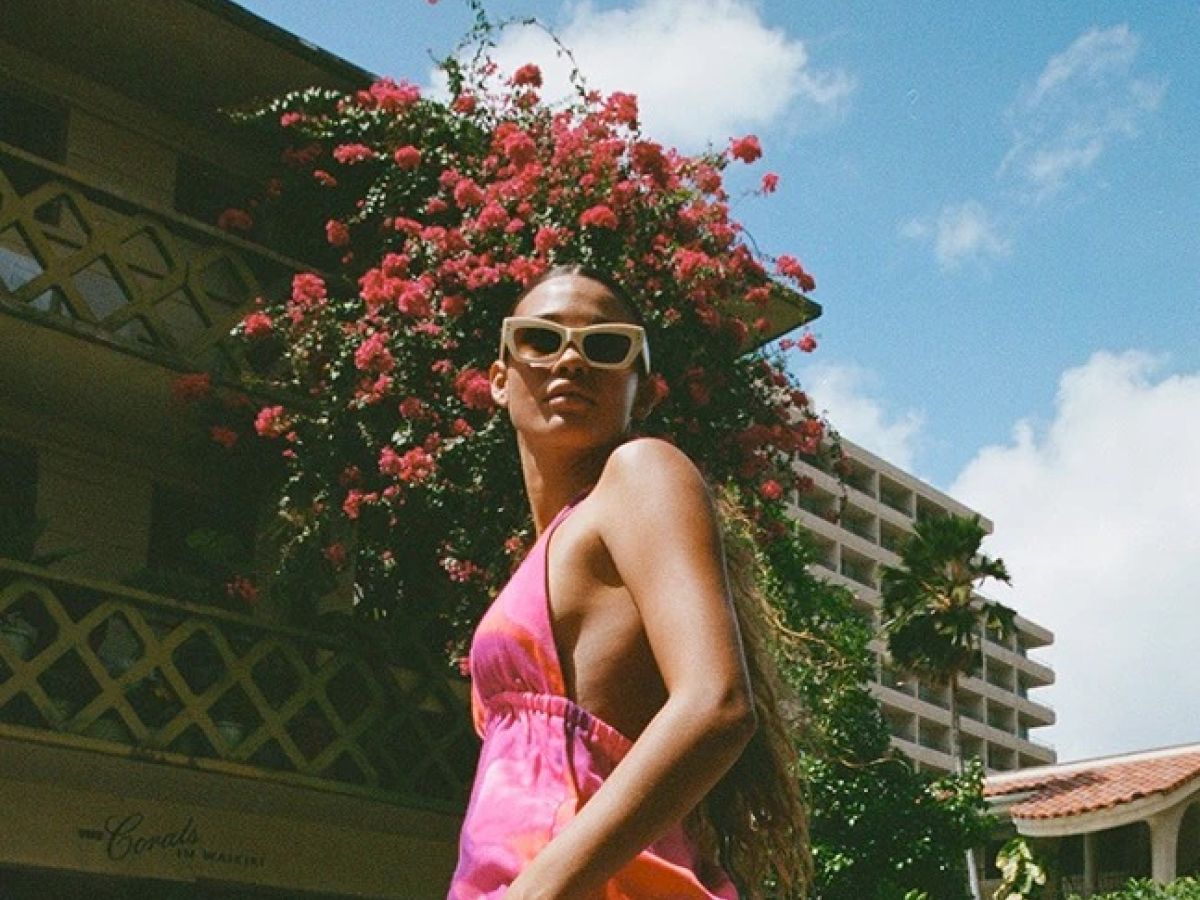 A person wearing a pink dress and sunglasses stands outdoors with a building and blooming flowers in the background under a partly cloudy sky.