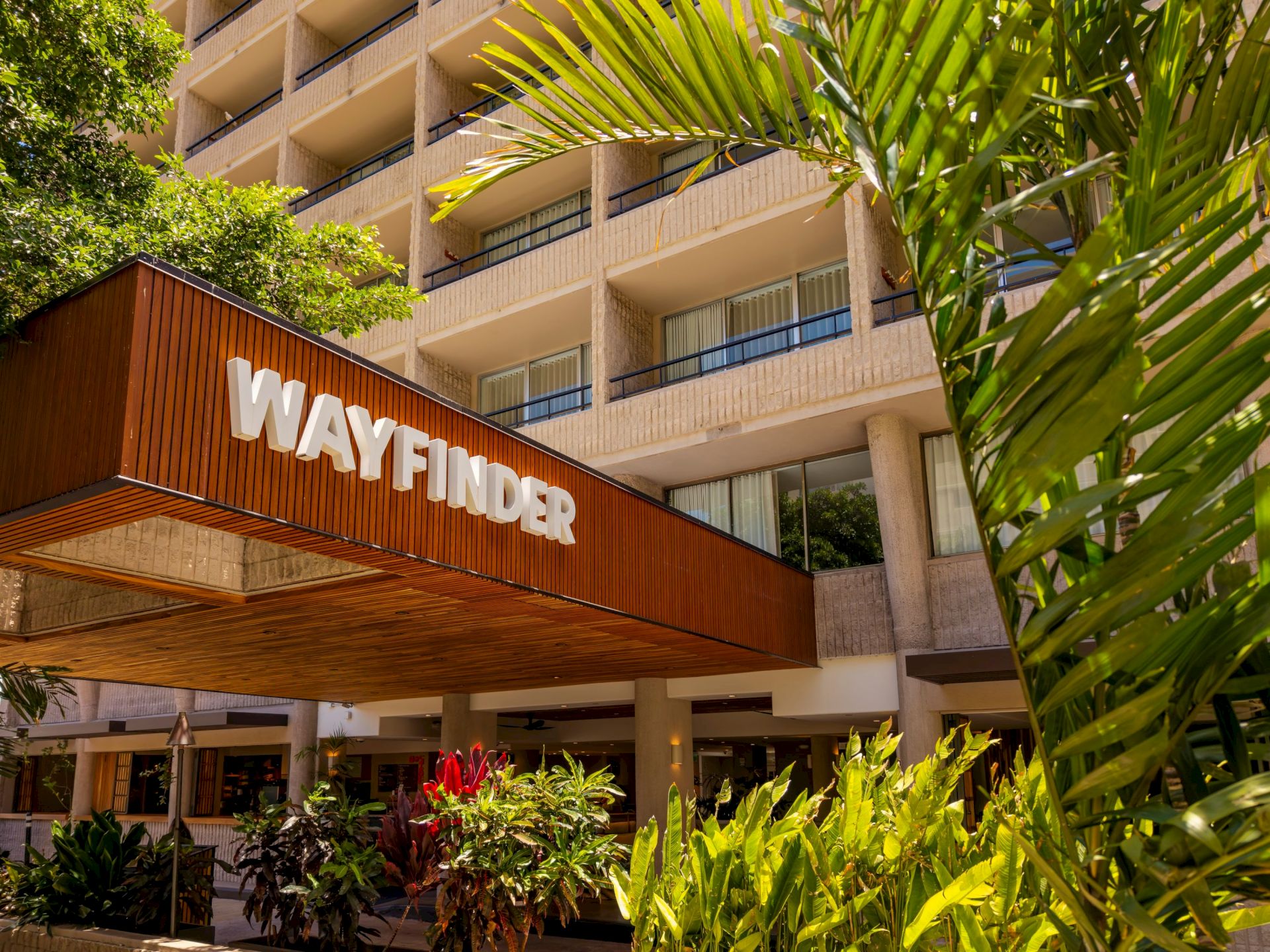 Se&ntilde;alizaci&oacute;n en la entrada de un edificio con plantas tropicales y un hotel u oficina de varias plantas detr&aacute;s.