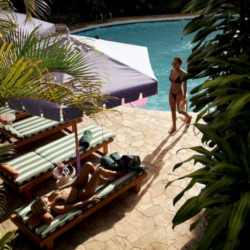 A sunny poolside scene with a woman walking beside the pool and two people relaxing on lounge chairs under umbrellas, surrounded by lush greenery.