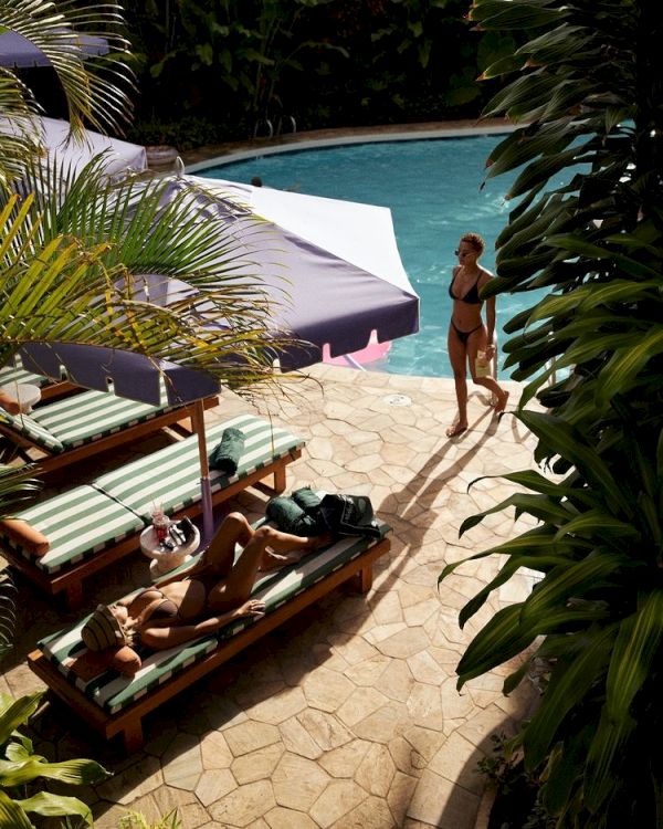 A sunny poolside scene with a woman walking beside the pool and two people relaxing on lounge chairs under umbrellas, surrounded by lush greenery.