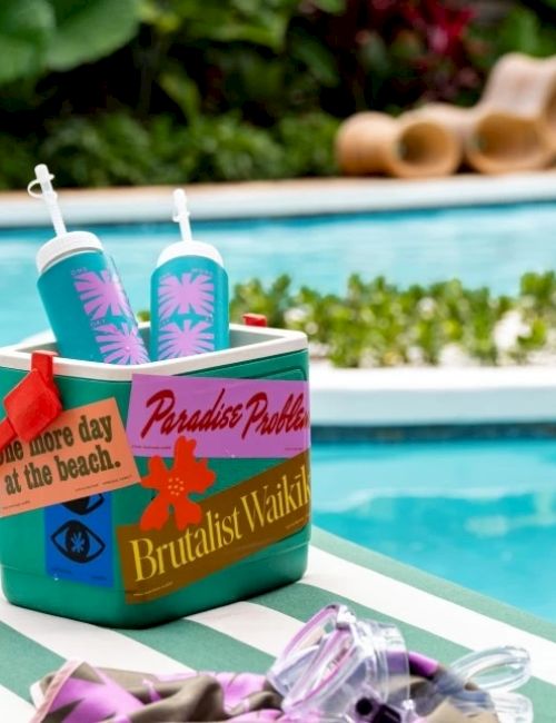 A cooler with decorative stickers and two teal water bottles near a pool. A pair of goggles and a towel are visible in the foreground.