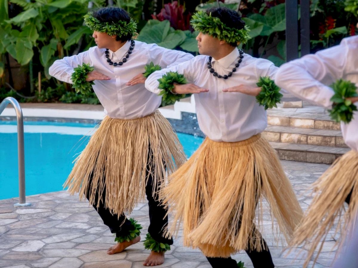 Dancers in traditional attire perform near a swimming pool, wearing grass skirts and greenery accessories, surrounded by lush plants.