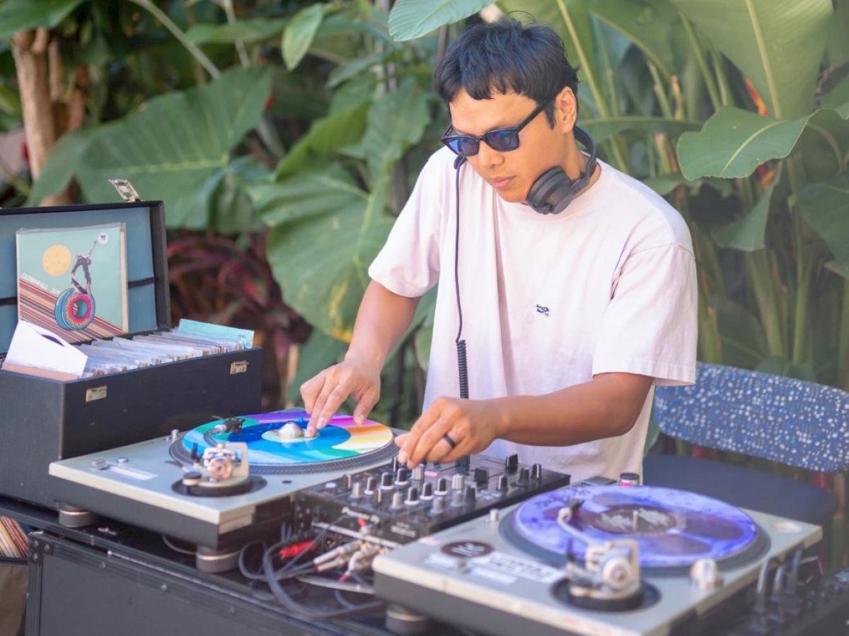 A DJ wearing sunglasses and a T-shirt is mixing music on a turntable setup outdoors, surrounded by lush green plants and trees ending the sentence.