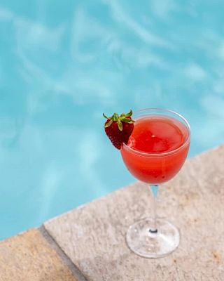 A glass of red drink garnished with a strawberry on the rim, placed on the edge of a pool.