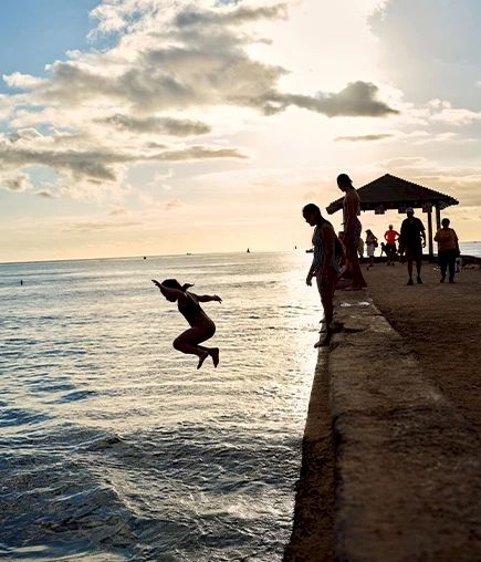 人々が桟橋に集まっている中、一人が水に飛び込む。背景には夕日が沈み、空には雲がいくつか浮かんでいる。