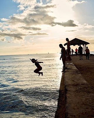 People are gathered on a pier as one person jumps into the water, with the sun setting in the background and a few clouds in the sky.