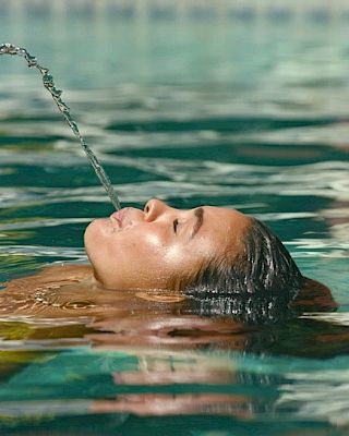 A person is floating in a pool with their head tilted back, spouting a stream of water from their mouth.