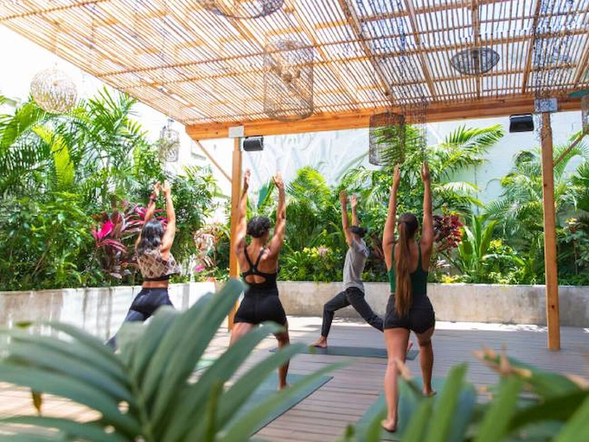 A group of people practicing yoga under a wooden canopy in an outdoor setting surrounded by lush greenery, with some raising their arms.