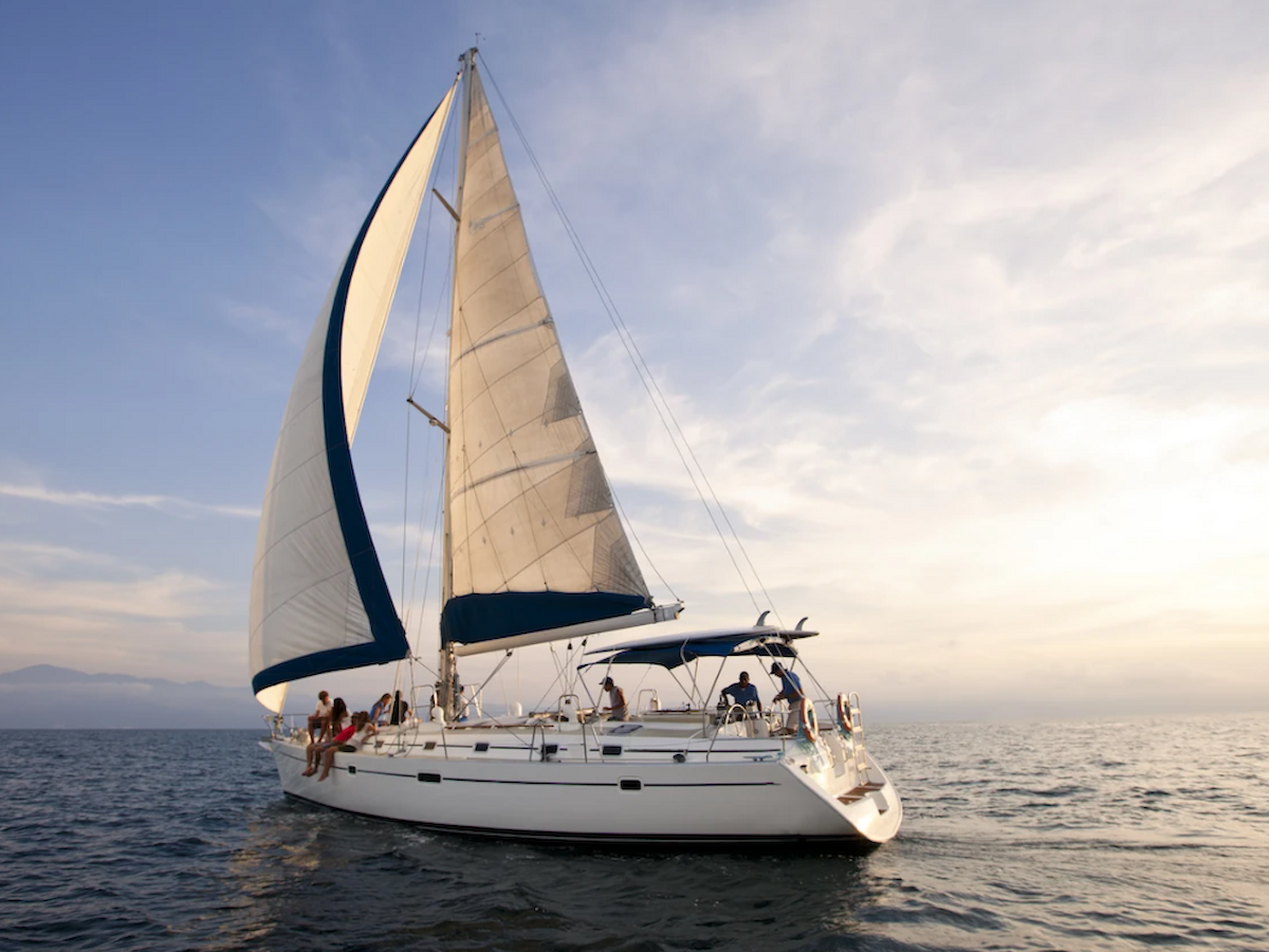 A sailboat with white sails is on a calm sea at sunset. People are on board, enjoying the view and the peaceful surroundings.