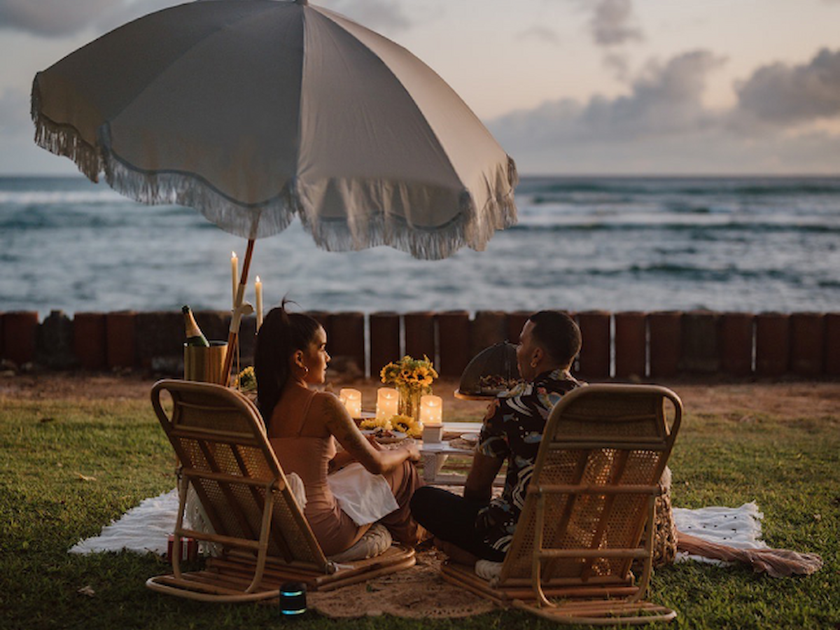 A couple is enjoying a beach picnic under an umbrella with candles and flowers on a blanket, with the ocean view in the background.