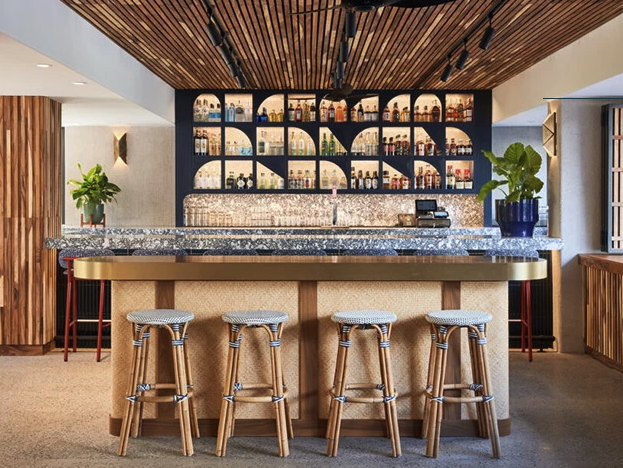The image shows a modern bar area with a marble countertop, four bar stools, plants, and a stylish back wall with shelves displaying bottles.