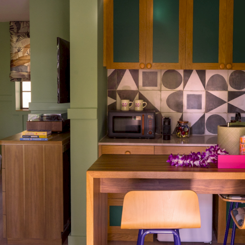 A modern kitchen with wooden cabinets, a geometric tile backsplash, a countertop of appliances, cups, and purple flowers, in daylight.