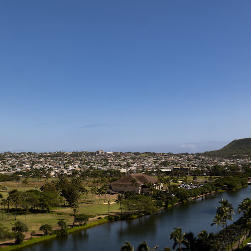 The image shows a river or canal running through a green landscape with city buildings in the background and a mountain under a clear blue sky.