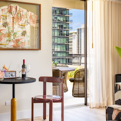 A room with tropical decor: framed shirt, table with wine, chairs, plant, sofa, balcony view with buildings.