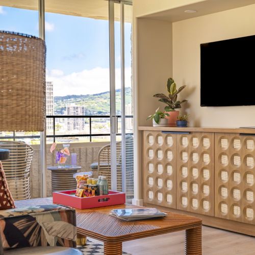 A cozy living room with a TV, cabinet, and plants. A balcony view with seating and a mountain backdrop completes the inviting atmosphere.