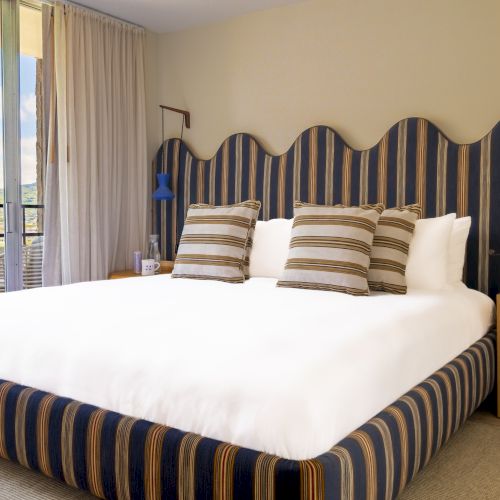 The image shows a stylish bedroom with a striped bed, pillows, wooden nightstands, and a balcony view of a cityscape under a bright sky.