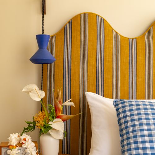 A cozy bedroom with a striped headboard, checkered pillow, wooden side table with a flower arrangement, and a hanging blue lamp.