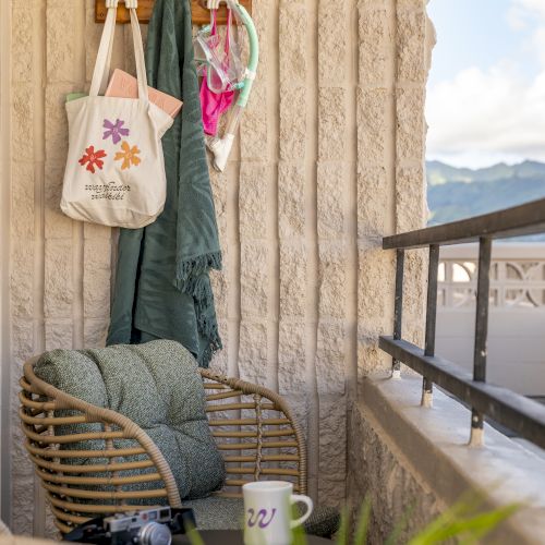 A cozy balcony with rattan chairs, a mug, a bag, and a jacket on hooks. The view includes mountains under a clear sky.