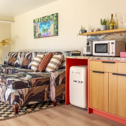 A cozy room features a floral sofa, a checkered rug, a kitchenette with a fridge and microwave, and a vibrant wall art piece.