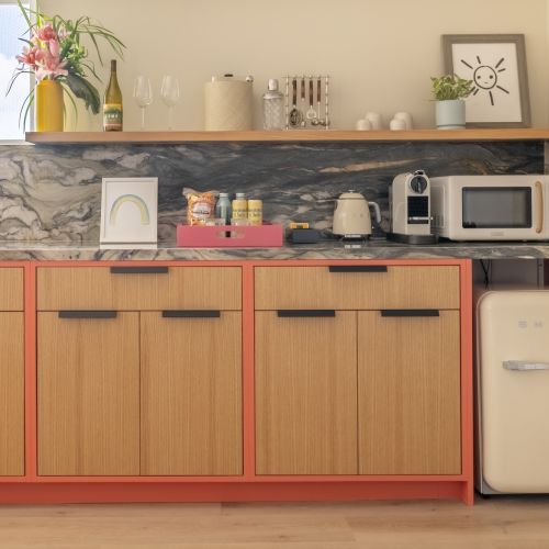 A modern kitchen with wooden cabinets, marble backsplash, microwave, toaster, kettle, and a small refrigerator. A vase with flowers is on top.